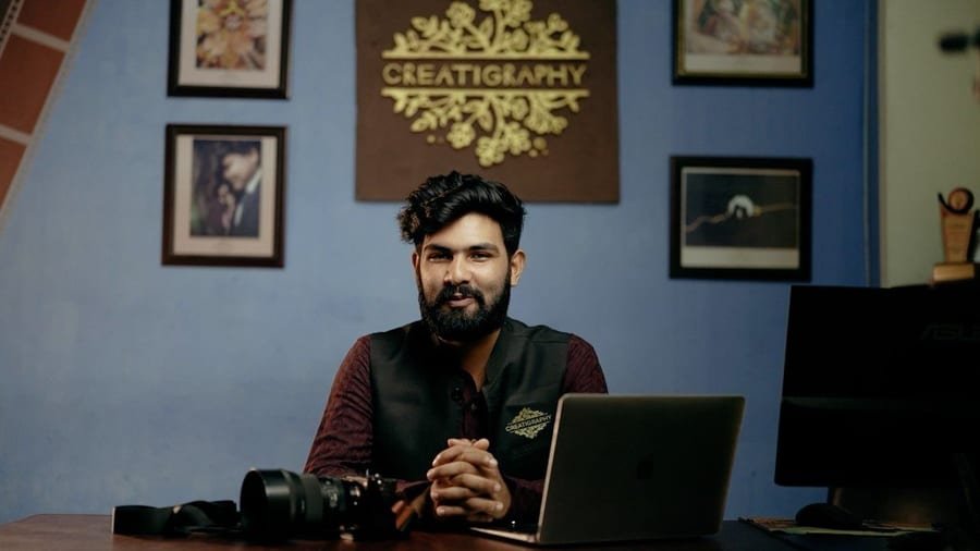 A member of the Creatigraphy team at the studio in Khoksa, Kushtia, with framed prints and the Creatigraphy wall sign behind him